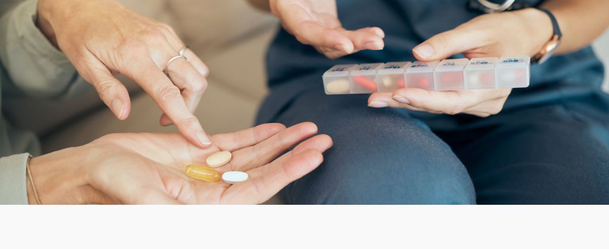 Eine ältere Frau hält mehrere Medikamnete in der Hand und eine Apothekerin zeigt mit Ihrer Hand auf eine Medikamnetenbox